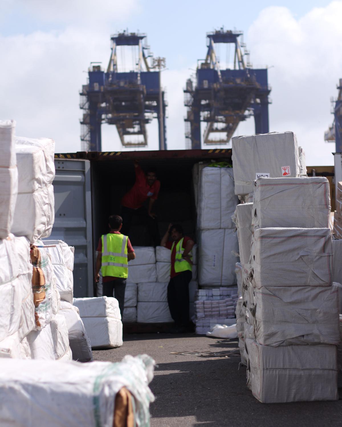 Bales of goods beside an open shipping container at a port, with two cranes behind
